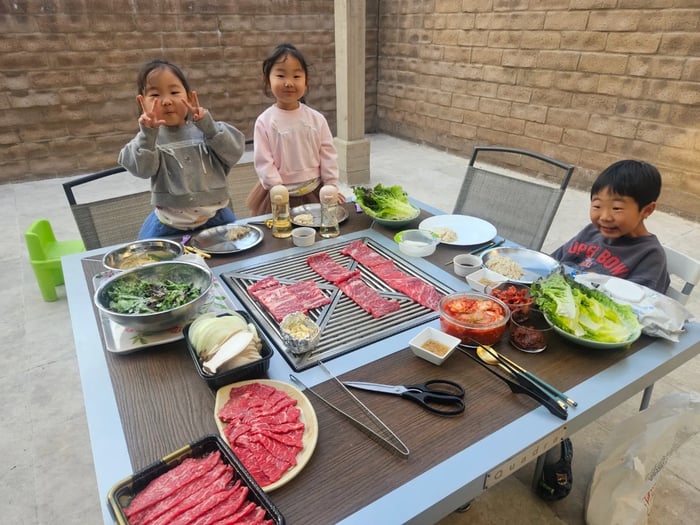  A candid photo shows three children sitting at a BBQ grill table on a patio, ready for a meal. The table is filled with plates of thinly sliced raw meat, side dishes like kimchi and salads, and grilling utensils