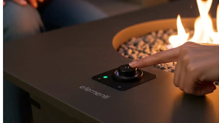 A hand pressing the ignition button on an Elementi fire pit table, showcasing the easy-to-use control panel and creating a warm and inviting atmosphere.