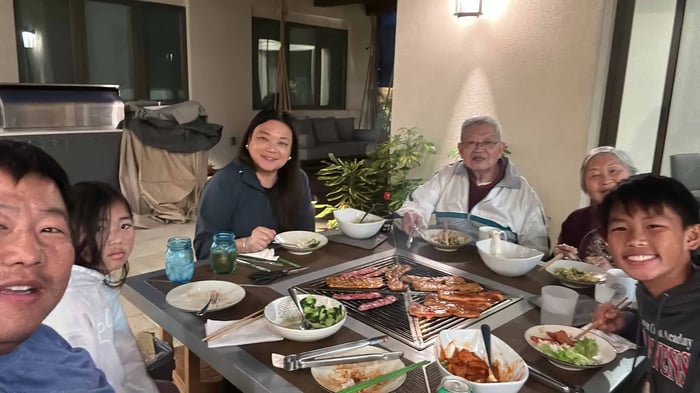  A multigenerational Asian family is gathered around a bbq grill table for an evening meal. The family is sitting closely together, smiling at the camera as they eat. On the grill in the center of the table, various meats and sausages are cooking.