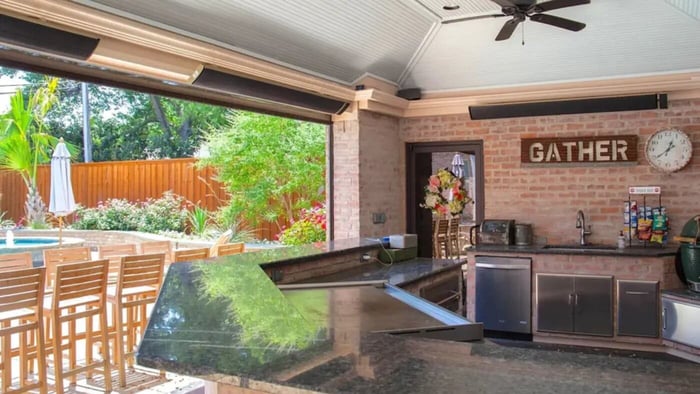A luxurious outdoor kitchen and bar area featuring a brick wall, ceiling fan, and two black wall-mounted electric patio heaters installed over the counter and seating area.