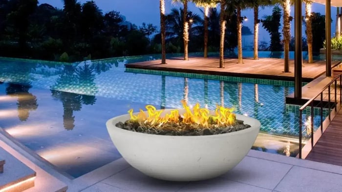 A white, rounded concrete fire bowl filled with dark rocks and flames, positioned near a multi-level modern swimming pool with a wooden deck and thin trees at dusk.