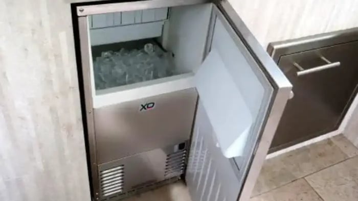 Stainless steel under-the-counter ice maker built into an outdoor kitchen. The door is open, revealing a bin full of ice cubes.