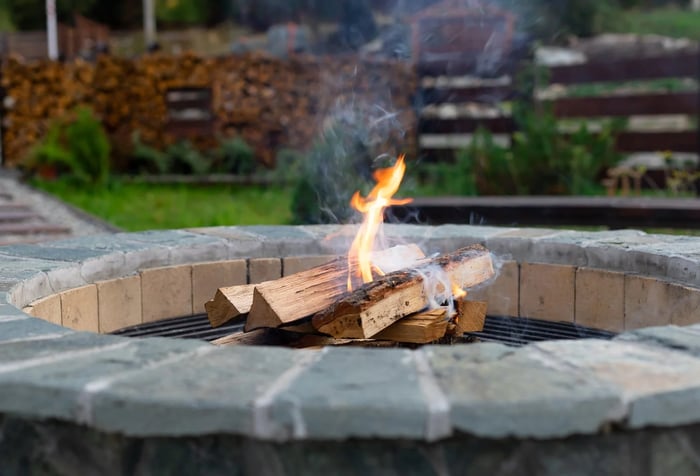 A brick fire pit with a small fire burning inside. The fire pit is surrounded by a lush green lawn and a wooden fence.