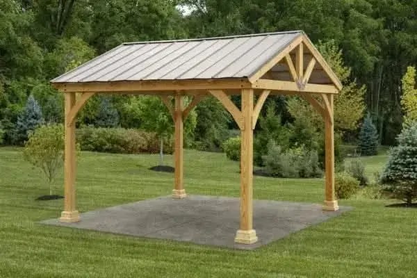 A light-colored wooden a frame pavilion with a gray metal roof, resting on a square concrete pad in a green, grassy outdoor setting.