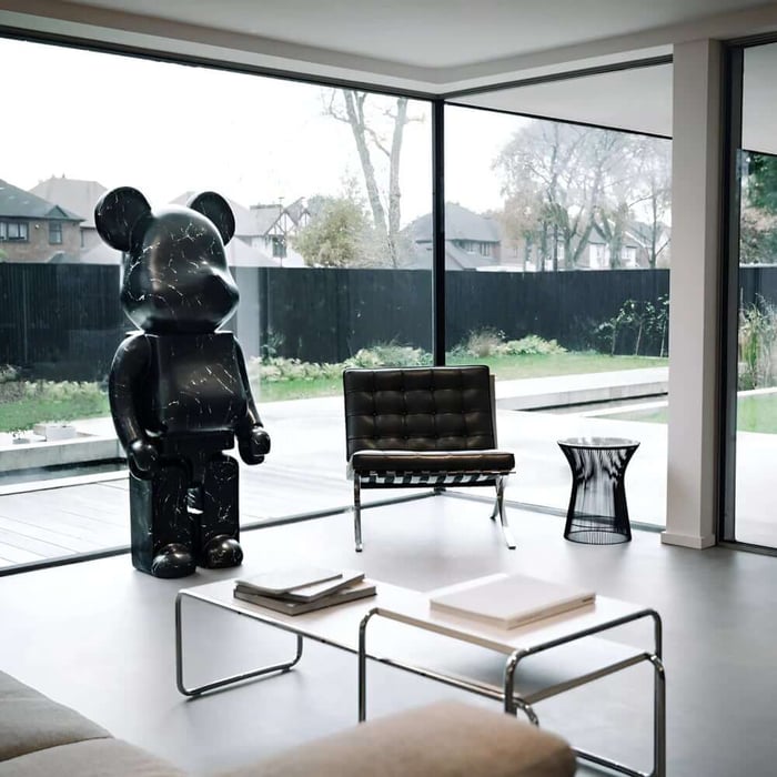 Modern living room with floor-to-ceiling corner windows, a large black bear-shaped sculpture by the glass, a black lounge chair, and a low metal coffee table with books.