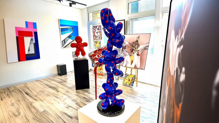 Blue stacked bear sculpture with red heart spots on a black base inside a modern art gallery, surrounded by vibrant pop-art paintings