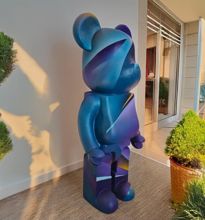 Large 165cm bear sculpture painted in a cosmic blue and purple gradient with abstract streaks of yellow and light blue, displayed on a terrace with natural sunlight and potted plants.