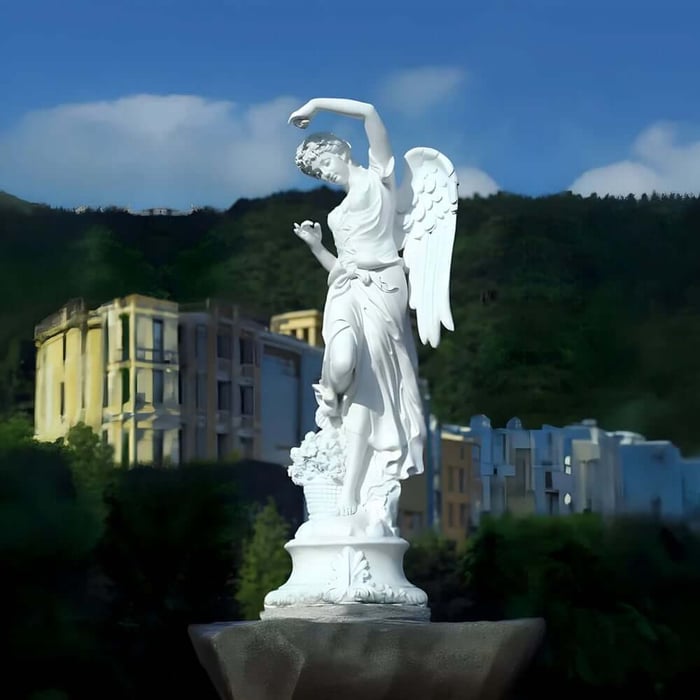 White angel marble sculptures statue with raised arm and wings on a pedestal, set against hills and buildings under a blue sky.