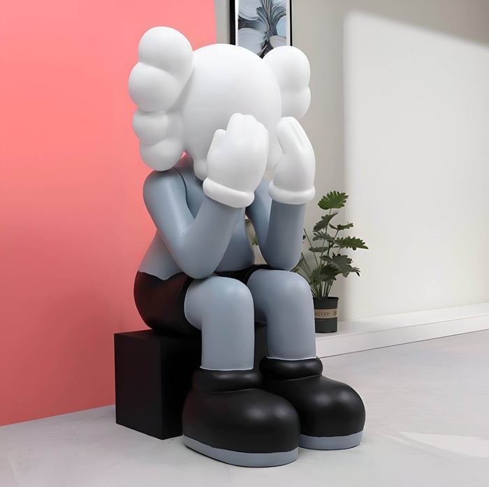 Large iconify sitting sculpture in slate grey and white, featuring a cartoon-like figure with gloved hands covering its face, displayed indoors against a pink and white wall with decorative plants.