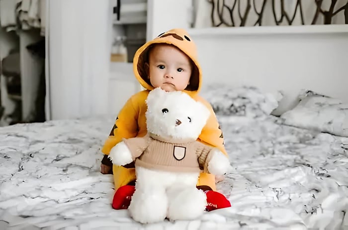 a baby wearing an orange baby suit sat on a grey bed with a white teddy bear in a brown jumper.