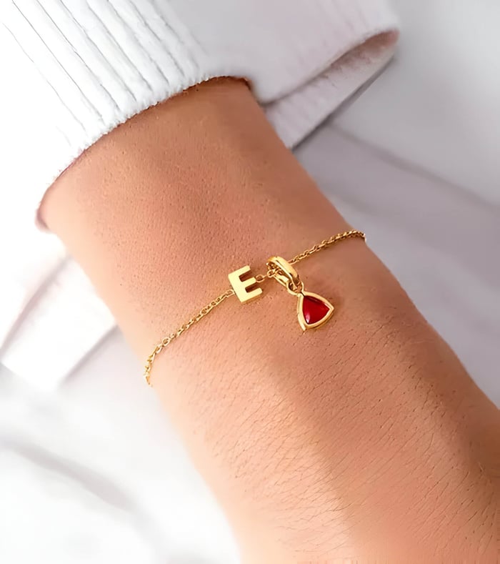 A gold bracelet with a red gem & a letter E, worn on someones wrist