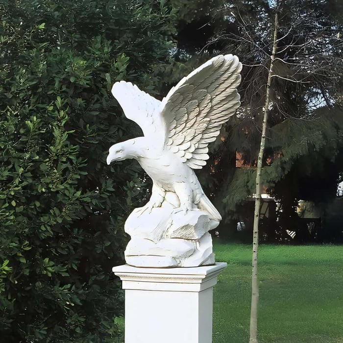 White eagle sculpture with wings spread, perched on a pedestal in a garden setting.