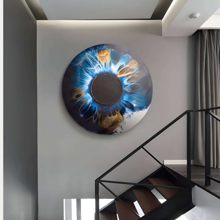 A modern staircase with wooden steps and a black railing ascends, and on the gray wall, the Genesis Earth Tones 3D Resin Crystal Porcelain LED Wall Art by Giant Sculptures displays swirling blue, black, and gold patterns resembling an abstract eye. Earth tones blend beautifully as natural light partially illuminates the space.