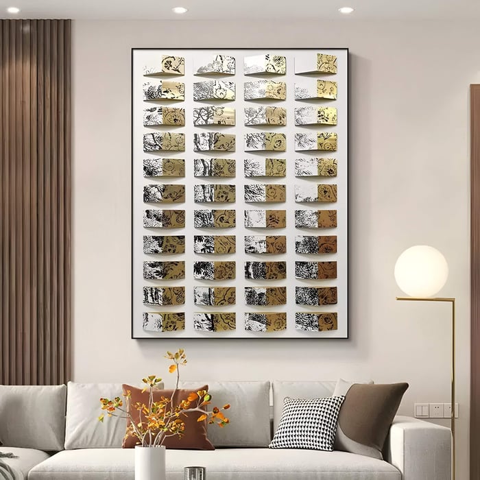 A contemporary living room features the Muraluxe Gold & Black Curved Mosaic 3D Wood Art by Giant Sculptures, composed of rows of rectangular panels. Below, a light gray sofa with white and gray cushions is paired with a round lamp and floral arrangement to complete the setting.