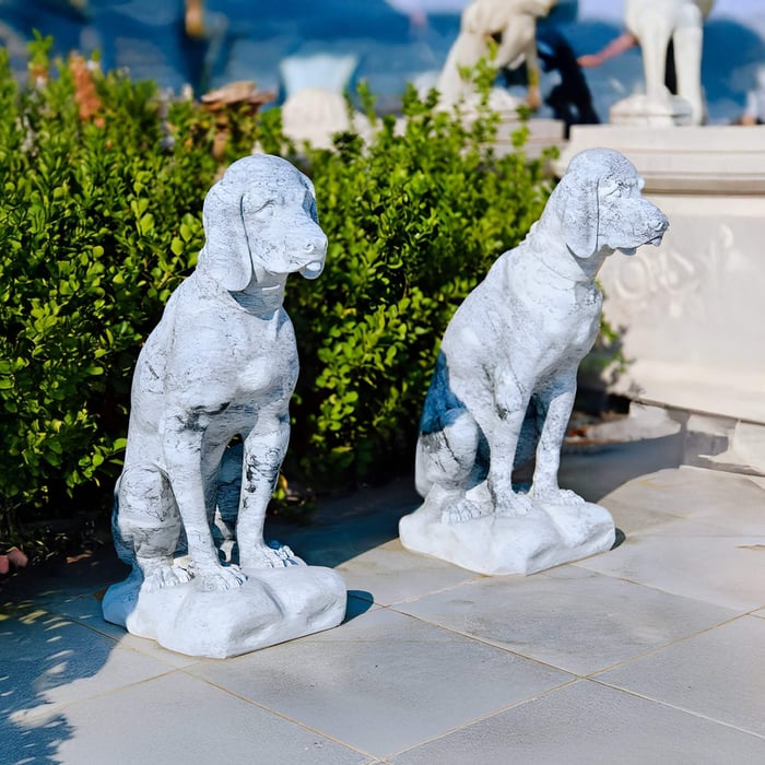 Twin marble dog statues seated side by side near shrubbery, adding charm and symmetry to a paved outdoor garden area.