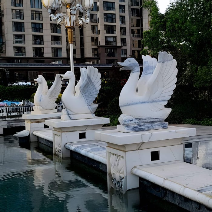 Giant Sculptures Wings-Lifted Swan II Marble Outdoor Sculptures, each 120cm tall, rest gracefully on square pedestals by a reflective pool. Behind them stands a modern apartment building with trees, and above is an elegant decorative lamppost.