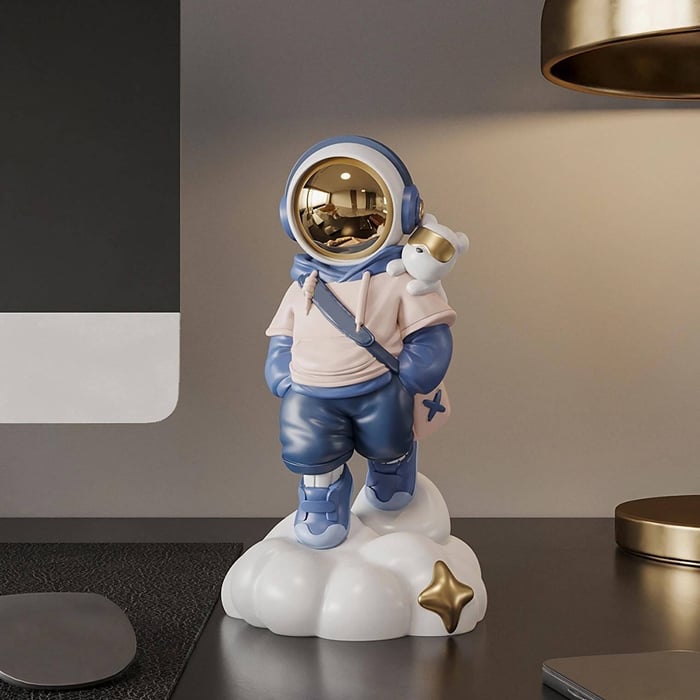Playful astronaut figurine with a gold visor, hoodie and crossbody bag, carrying a tiny bear on its shoulder, standing on a white cloud base with a gold star on a modern desk.