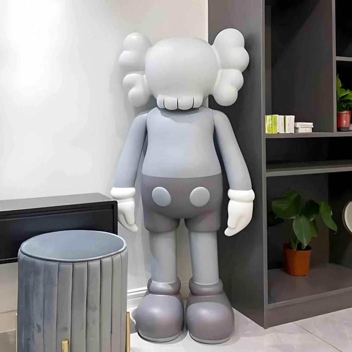 A large gray cartoon-style sculpture with rounded shapes and gloved hands stands in the corner of a modern room beside shelving and a small plant.