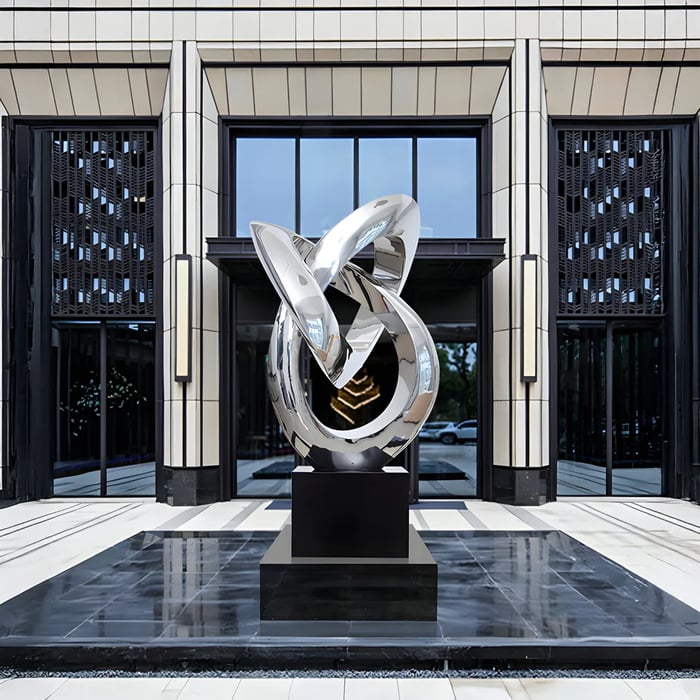 Large polished silver abstract looped sculpture on a black pedestal in front of a modern glass-and-tile building entrance, reflecting the surrounding plaza and sky.