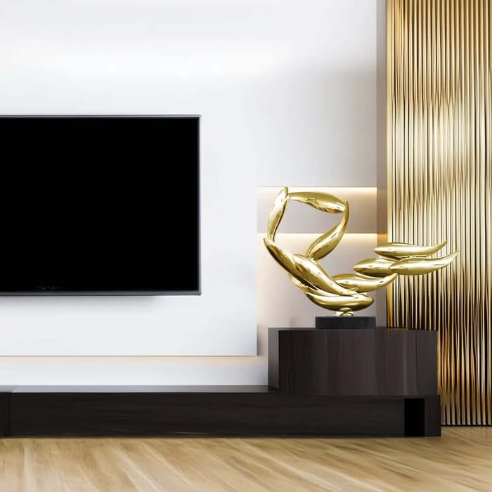 Gold abstract ribbon-like sculpture on a dark wood media console beside a flat-screen TV in a modern living room with white walls and a gold textured panel.