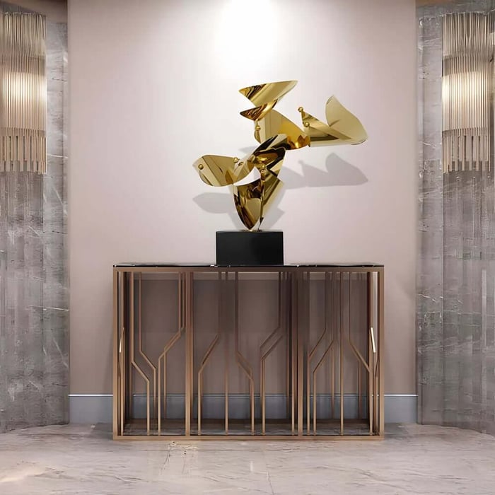 Gold abstract metal sculpture on a black plinth, centred on a console table in a minimalist interior setting.
