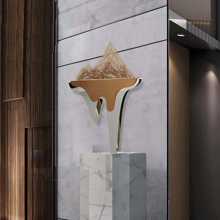 Gold mountain-shaped abstract sculpture on a marble pedestal in a modern interior.