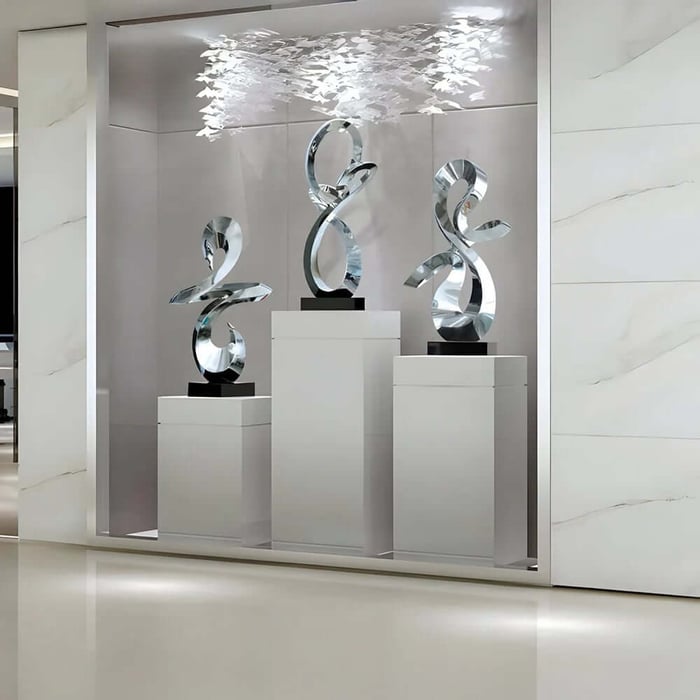 Three tall silver abstract ribbon sculptures on white pedestals in a recessed, modern display nook with soft overhead lighting and a cluster of delicate white hanging elements above.