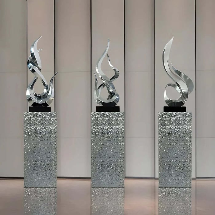 Three reflective silver abstract sculptures on textured stone pedestals, arranged in a clean gallery-style space.