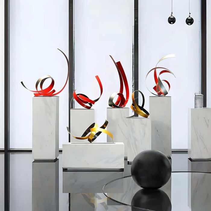 Series of ribbon-like abstract metal sculptures in red, gold, and black on white plinths by tall windows.