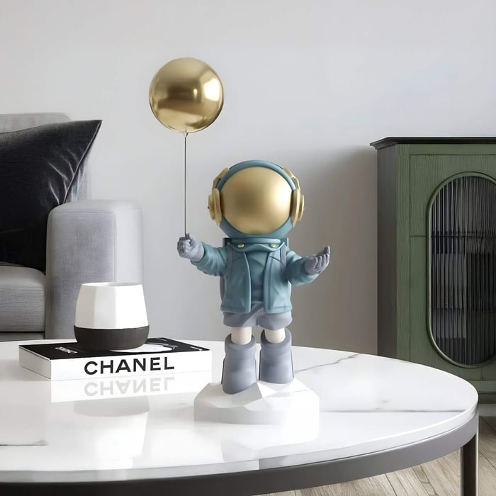 Blue astronaut sculpture with a gold visor holding a gold balloon on a geometric base, displayed on a white marble coffee table in a modern living room.