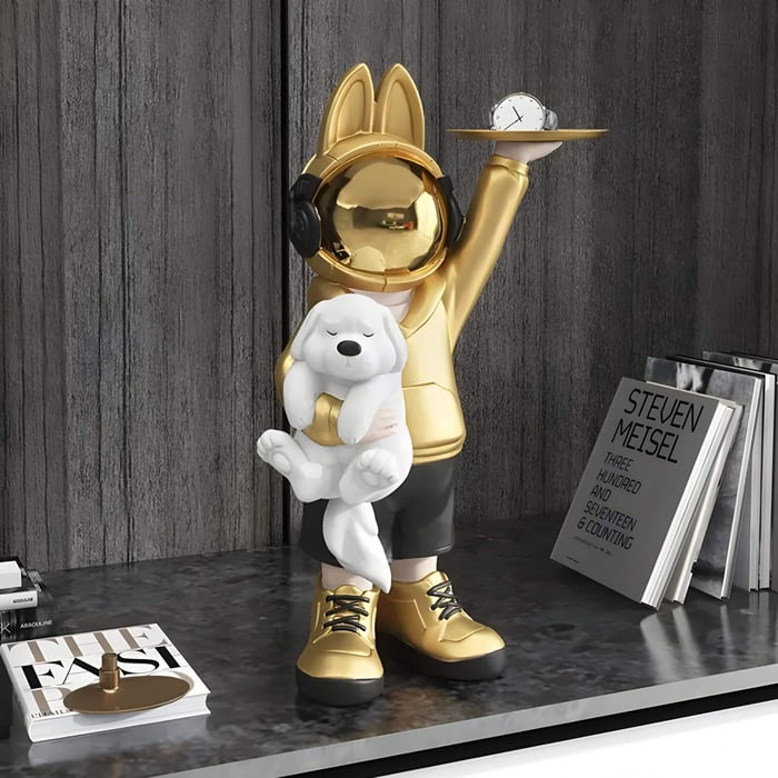 The Gold Astronaut Bunny Pup Butler Figure - 44cm by Giant Sculptures features a golden, bunny-eared statue with headphones holding a white dog and clock. Its displayed on a shelf with books, like STEVEN MEISEL, set against a wooden backdrop.