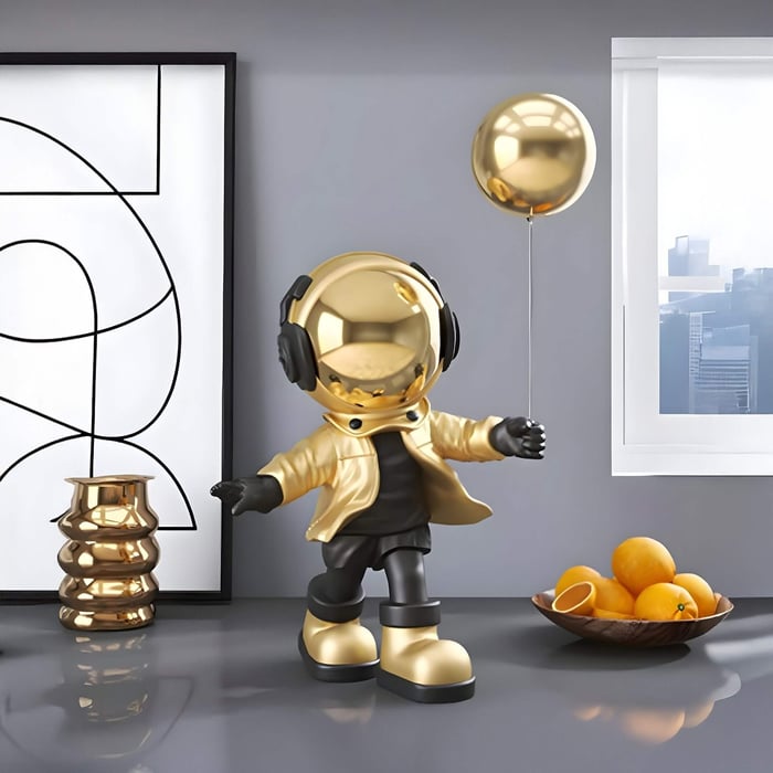Gold-and-black astronaut figurine with headphones and a gold jacket holding a gold balloon, standing on a glossy gray surface beside abstract art and a bowl of oranges near a window.