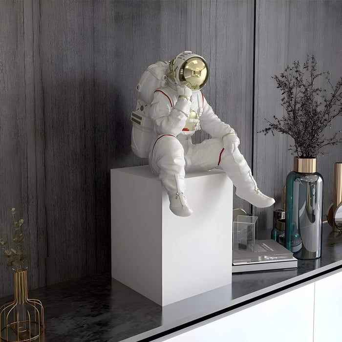 White astronaut sculpture with a gold visor in a thinker pose, seated on a white pedestal in a modern interior with gray walls, decorative vases, and books.