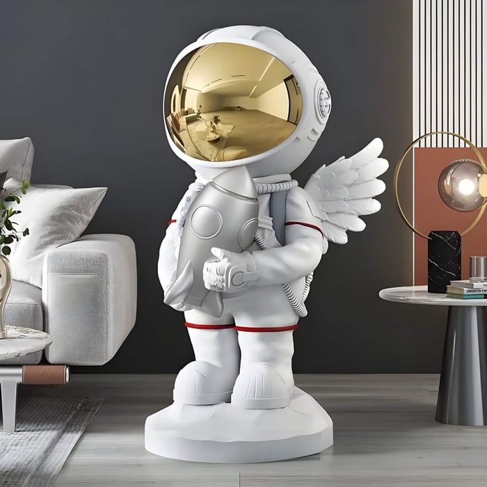 White astronaut sculpture with a gold visor and wings holding a rocket, standing on a pedestal in a modern living room next to a sofa and side table.