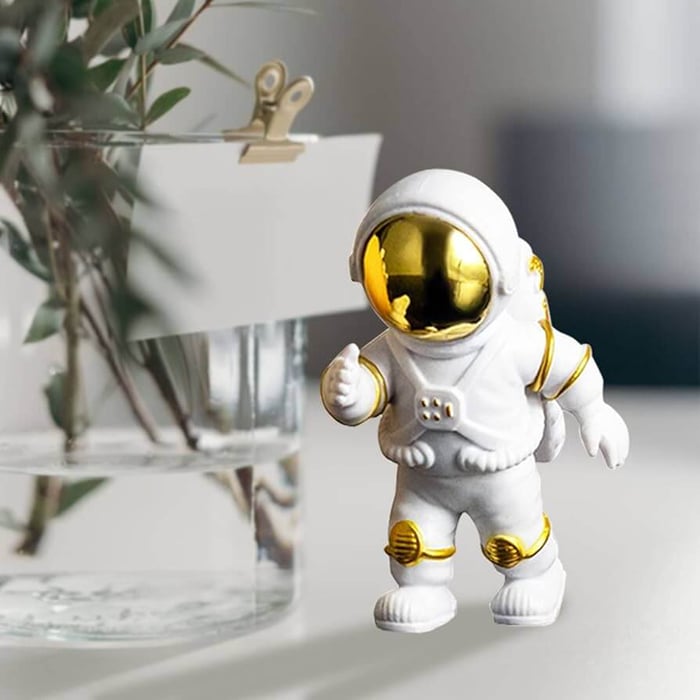 Small white astronaut figurine with golden visor and details standing on a table beside a glass vase.