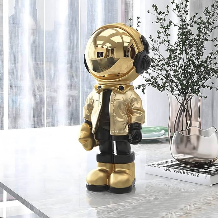 Gold astronaut figurine with black outfit and reflective helmet standing on a marble table beside a vase and decorative books.