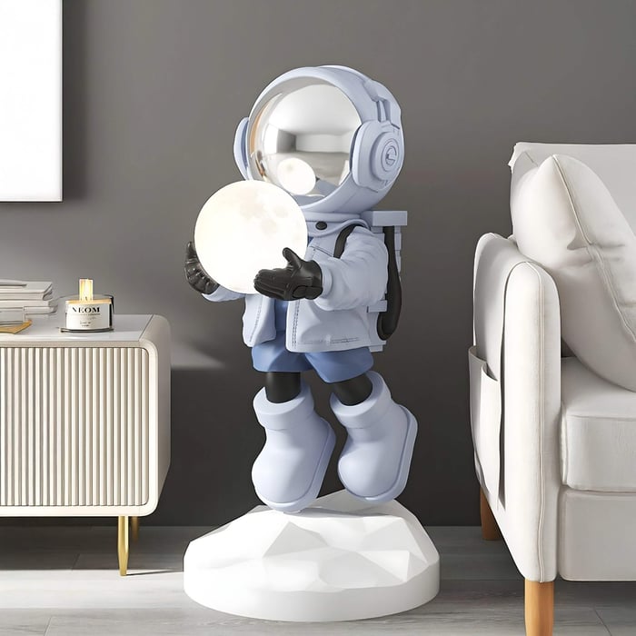 The Giant Sculptures Cosmic Sky Astronaut Glow Hover Sculpture - 120cm features a reflective helmet and large boots on a white base, holding a glowing moon. It stands beside a modern beige sofa and white cabinet with a candle, against a gray wall.