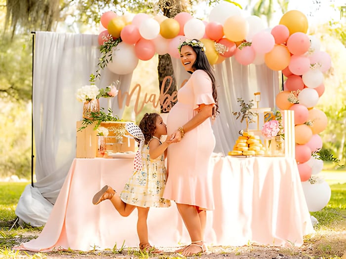 A woman at a baby shower with decorations behind, and child kissing her bump
