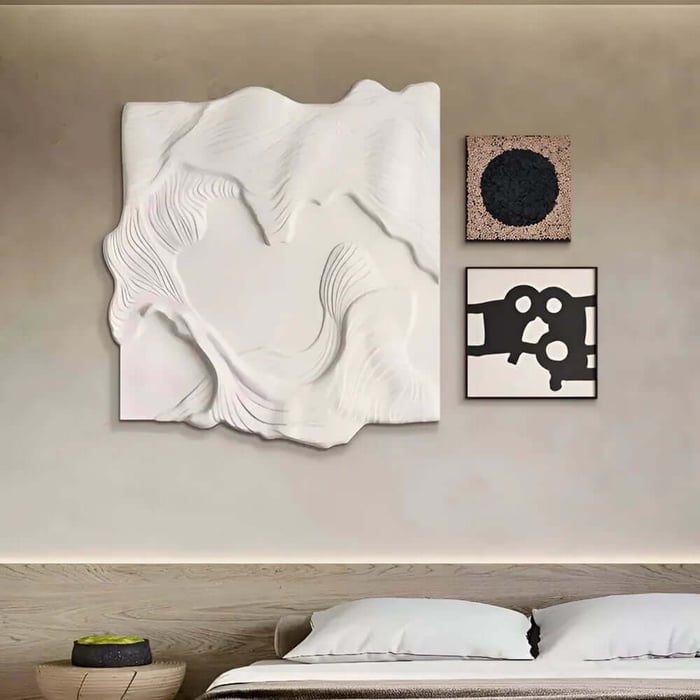 Minimalist bedroom with a light wood headboard and three-piece wall art arrangement above the bed, featuring a large white sculpted relief panel, a small cork panel with a black circle, and a black-and-white abstract print.