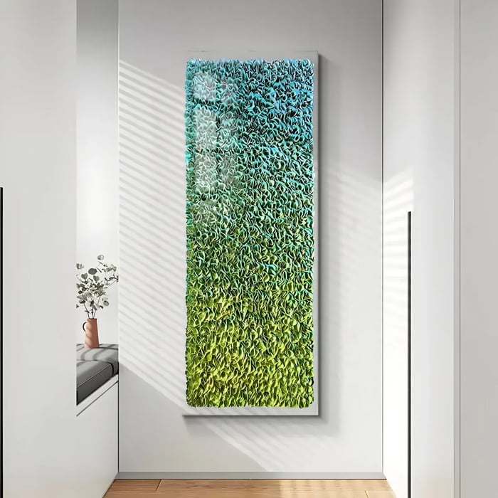 A tall, vertical wall panel with a three-dimensional textured surface transitioning from blue to green, mounted in a bright hallway with natural light streaming in. A built-in bench and vase with flowers complement the modern setting.