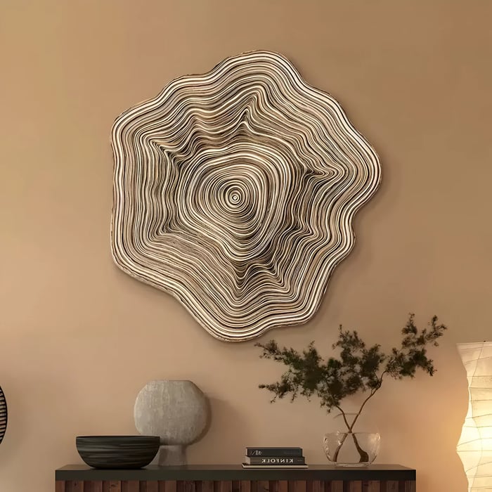 A large sculptural wall art piece resembling the cross-section of a tree trunk, with intricate concentric layers mimicking natural wood grain. Mounted on a warm-toned wall above a wooden sideboard, the setting is complemented by neutral decorative elements, including ceramic vases, a small plant in a glass container, and soft ambient lighting, creating a cozy and organic atmosphere.
