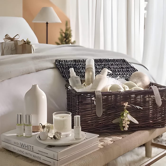 a brown wicker hamper filed with The White Co. spa gifts, placed on a table at the end of a bed as Christmas Gifts for In-Laws