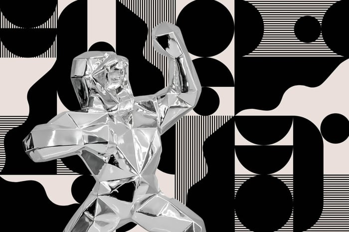 Polished silver geometric gorilla sculpture with angular faceted surfaces, posed with raised fists against a black-and-white abstract geometric background.