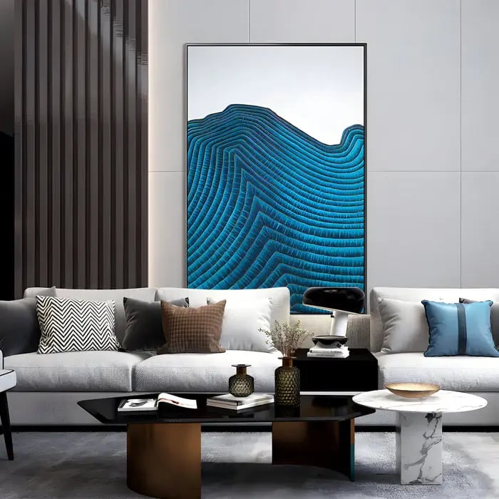A modern living room featuring a large abstract wall art piece with a textured blue topographic design, framed in black and mounted above a stylish grey sofa adorned with patterned and solid-colored cushions. 