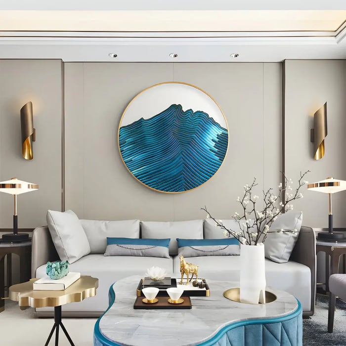 A modern and stylish living room featuring a circular abstract wave wall art piece in shades of blue, creating a focal point above a comfortable sofa with light blue and teal cushions and gold accent tables.