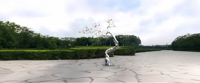 Modern garden sculptures of a stylized tree with a twisting metal trunk and delicate branches adorned with red blossoms, set in an outdoor plaza with lush green surroundings.