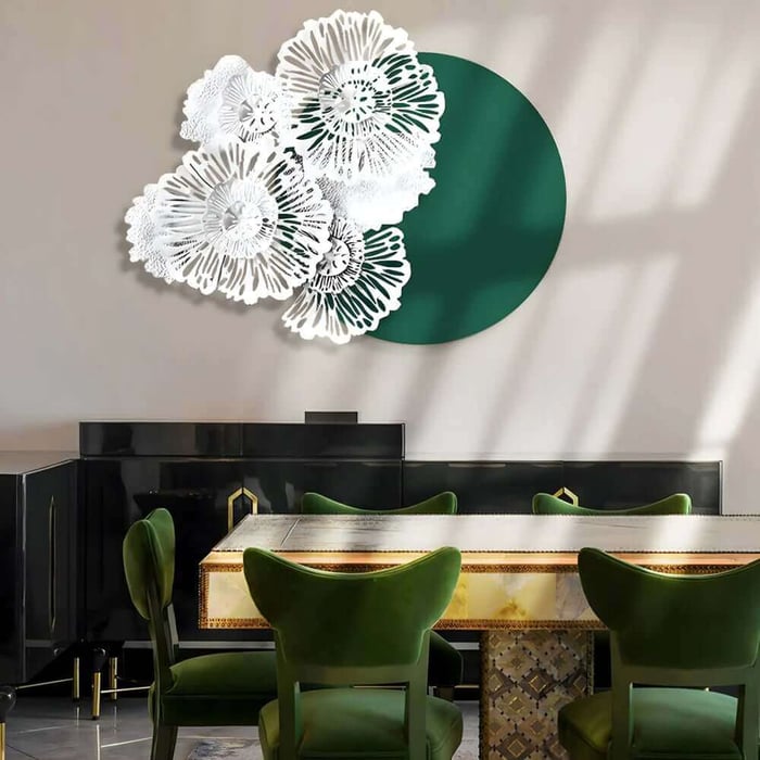 White floral metal wall sculpture layered over a large dark green circular panel, hanging above a luxurious dining table with green velvet chairs and black cabinetry in a sunlit room.
