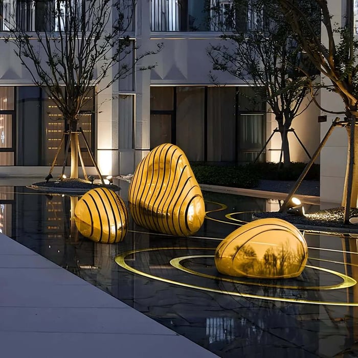 Three smooth golden abstract steel pebble sculptures installed in a reflective water courtyard at dusk.