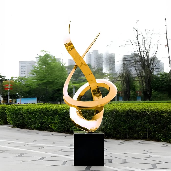 Golden abstract looping sculpture in a city plaza – A twisting gold metal sculpture standing on a black pedestal in an open urban space.