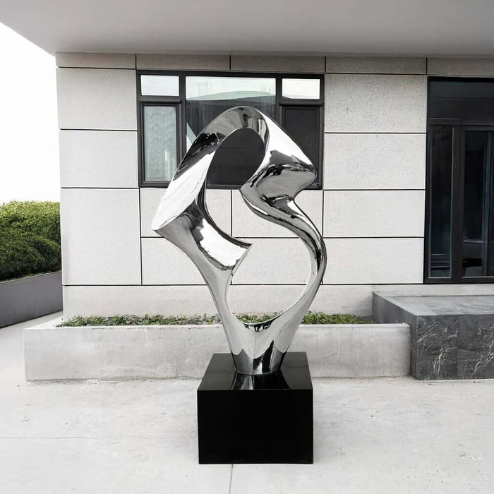 Polished silver abstract metal sculpture with a looping, hollow center on a black pedestal, displayed on a concrete patio in front of a modern building with light gray panel walls and dark-framed windows.